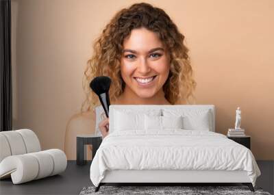 Young blonde woman with curly hair isolated on beige background holding makeup brush and whit happy expression Wall mural