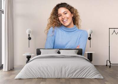 Young blonde woman with curly hair wearing a turtleneck sweater isolated on beige background keeping the arms crossed in frontal position Wall mural