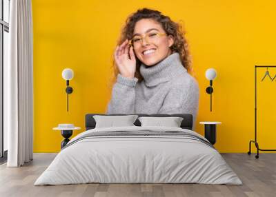 Young blonde woman with curly hair wearing a turtleneck sweater isolated on yellow background with glasses and happy Wall mural