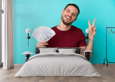 Young Brazilian man taking a lot of money over isolated background smiling and showing victory sign Wall mural