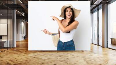 Young Brazilian woman with Pamela holding a beach bag isolated on white background surprised and pointing side Wall mural