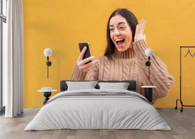 Young brunette woman at outdoors looking at the camera while using the mobile with surprised expression Wall mural