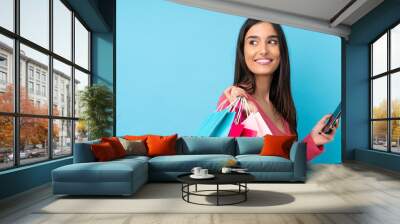 Young brunette woman over isolated blue background holding shopping bags and a mobile phone Wall mural