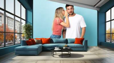 Young caucasian couple isolated on blue background whispering something Wall mural