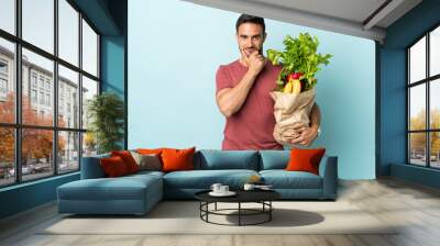 Young caucasian man buying some vegetables isolated on blue background laughing Wall mural