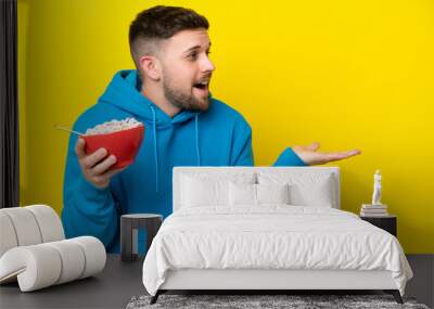 Young caucasian man holding a bowl of cereals isolated on yellow background with surprise facial expression Wall mural