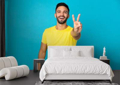 Young caucasian man isolated on blue background smiling and showing victory sign Wall mural