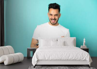 Young caucasian man isolated on blue background with phone in victory position Wall mural