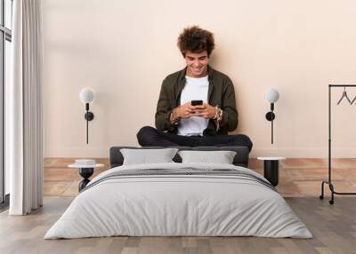 Young caucasian man sitting on the floor sending a message with the mobile Wall mural