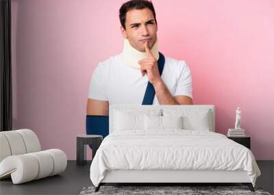 Young caucasian man with broken arm and wearing a sling isolated on pink background having doubts while looking up Wall mural