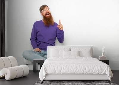 Young caucasian reddish man sitting on the floor isolated on white background pointing up and surprised Wall mural