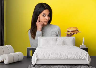 Young caucasian woman holding a burger isolated on yellow background thinking an idea Wall mural