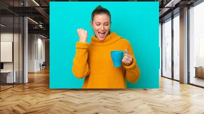 Young caucasian woman holding cup isolated on blue background celebrating a victory Wall mural