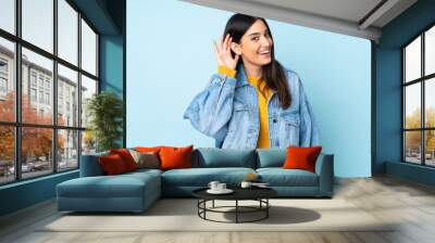 Young caucasian woman isolated on blue background listening to something by putting hand on the ear Wall mural