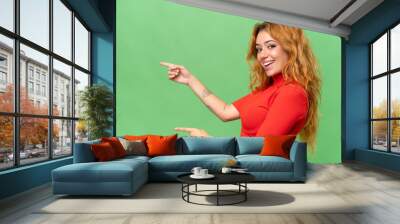 Young caucasian woman isolated on green screen chroma key background pointing finger to the side and presenting a product Wall mural