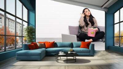 Young Colombian girl holding a bowl of popcorns and showing a film in a laptop making the gesture of madness putting finger on the head Wall mural