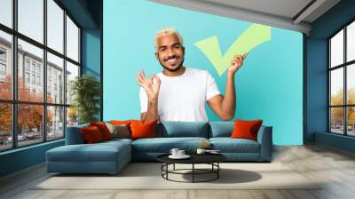 Young Colombian handsome man isolated on blue background holding a check icon and doing OK sign Wall mural