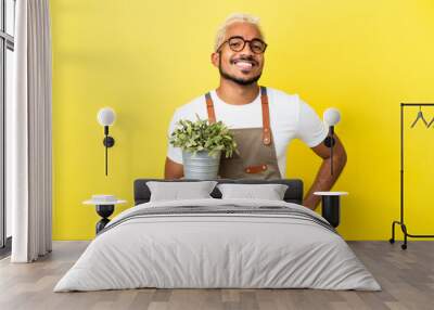 Young Colombian man holding a plant isolated on yellow background posing with arms at hip and smiling Wall mural