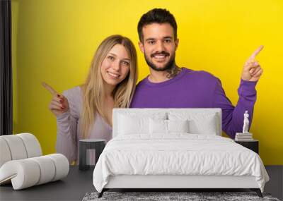 Young couple over isolated yellow background pointing finger to both sides Wall mural