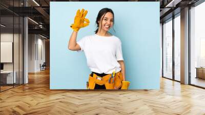 Young electrician woman isolated on blue background surprised and showing ok sign Wall mural