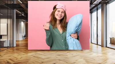 Young English woman in pajamas isolated on pink background in pajamas and inviting to come with hand. Happy that you came Wall mural