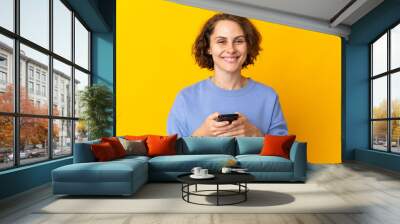 Young English woman isolated on pink background looking at the camera and smiling while using the mobile Wall mural