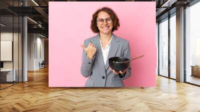 Young English woman isolated on pink background pointing to the side to present a product while holding a bowl of noodles with chopsticks Wall mural