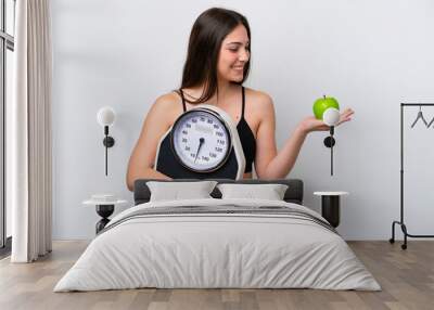 Young girl isolated on white background holding a weighing machine while looking an apple Wall mural