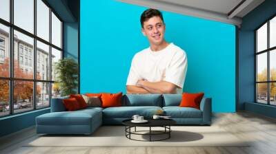 Young handsome Brazilian man isolated on blue background with arms crossed and happy Wall mural