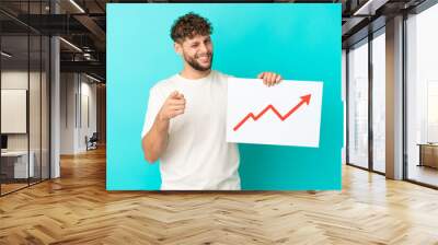 Young handsome caucasian man isolated on blue background holding a sign with a growing statistics arrow symbol and pointing to the front Wall mural