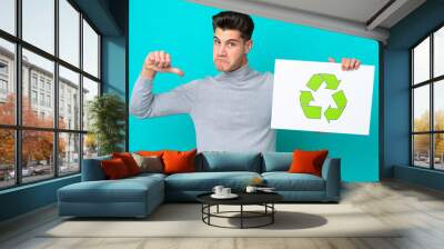 Young handsome caucasian man isolated on blue bakcground holding a placard with recycle icon with proud gesture Wall mural