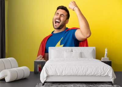 Young handsome caucasian man isolated on yellow background in superhero costume and celebrating a victory Wall mural