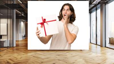 Young handsome man holding a gift over isolated background with surprise and shocked facial expression Wall mural