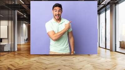 Young handsome man isolated on purple background surprised and pointing side Wall mural