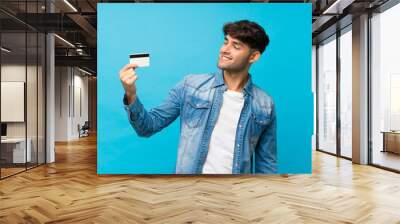 Young handsome man over isolated blue background holding a credit card Wall mural