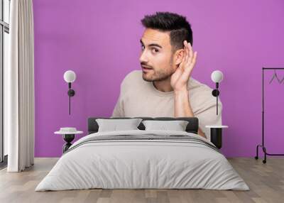 Young handsome man over isolated purple background listening to something by putting hand on the ear Wall mural