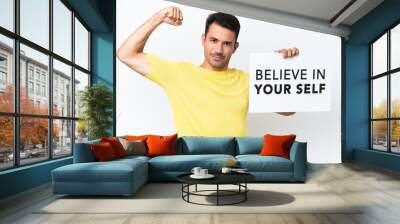 Young handsome man over isolated white background holding a placard with text Believe In Your Self and doing strong gesture Wall mural