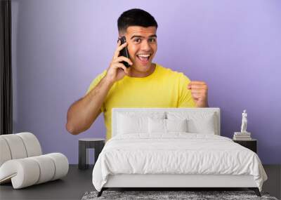 Young handsome man using mobile phone over isolated purple background celebrating a victory in winner position Wall mural