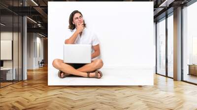 Young handsome man with a laptop sitting on the floor isolated on white background having doubts and with confuse face expression Wall mural