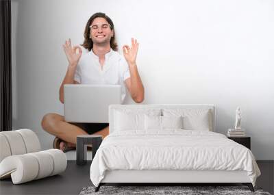 Young handsome man with a laptop sitting on the floor isolated on white background in zen pose Wall mural