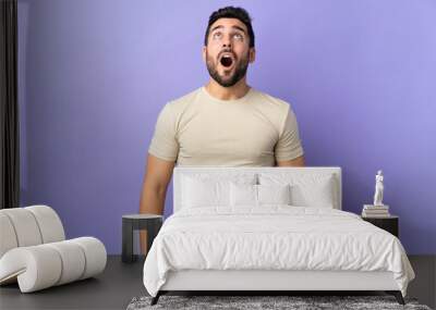 Young handsome man with beard over isolated background looking up and with surprised expression Wall mural