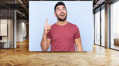 Young handsome man with beard over isolated blue background pointing up and surprised Wall mural