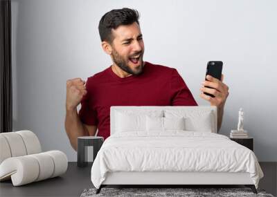 Young handsome man with beard using mobile phone isolated on white background celebrating a victory Wall mural