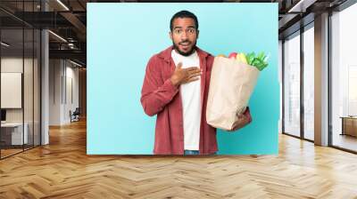 Young latin man holding a grocery shopping bag isolated on blue background surprised and shocked while looking right Wall mural