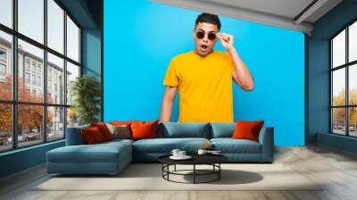 Young man with yellow shirt over isolated blue background with glasses and surprised Wall mural