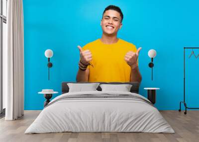 Young man with yellow shirt over isolated blue background with thumbs up gesture and smiling Wall mural