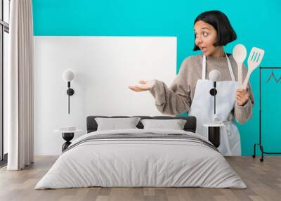 Young mixed race cook woman with a big placard isolated on blue background with surprise expression while looking side Wall mural