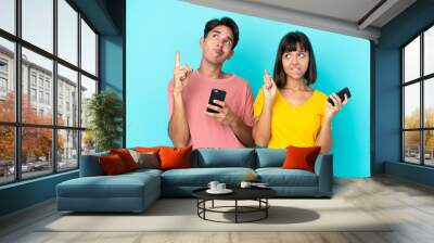 Young mixed race couple holding mobile phone isolated on blue background with fingers crossing and wishing the best Wall mural