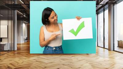 Young mixed race woman isolated on blue background holding a placard with text Green check mark icon and  pointing it Wall mural