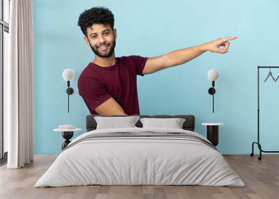 Young Moroccan man isolated on blue background pointing finger to the side and presenting a product Wall mural